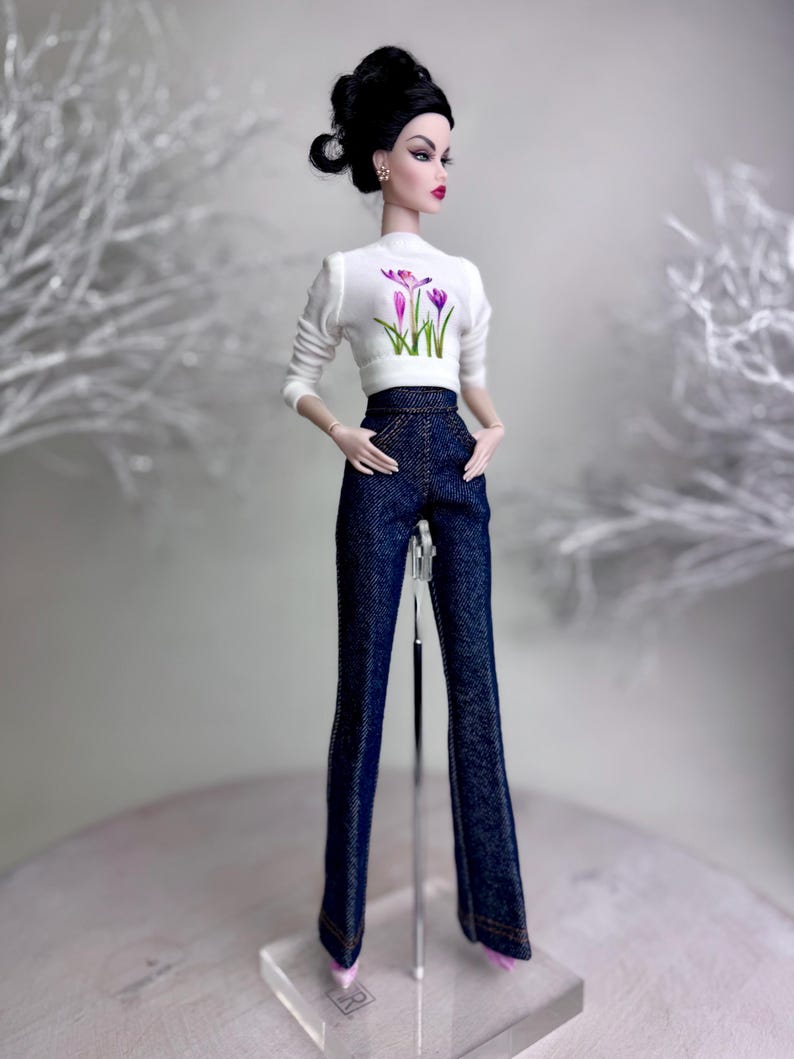 Op de afbeelding: Een modepop met een witte top met lange mouwen met een bloemenontwerp en een donkerblauwe spijkerbroek. De pop heeft zwart haar in een knot en oorbellen. De pop staat op een transparante standaard.
