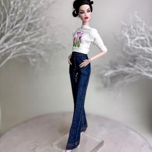 Op de afbeelding: Een modepop gekleed in een witte top met een bloemenprint en donkerblauwe jeans. De pop heeft donker haar in een updo en roze schoenen. De pop staat op een doorzichtige standaard. De jeans heeft een uitlopende pijp.