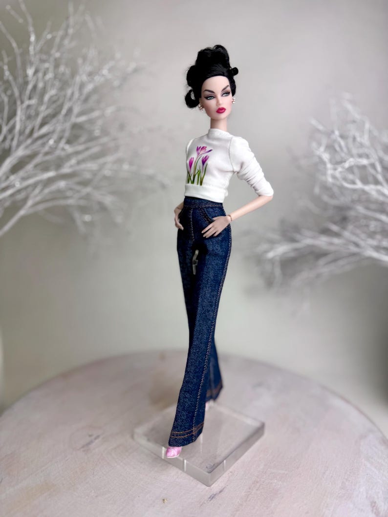 Op de afbeelding: Een modepop gekleed in een witte top met een bloemenprint en donkerblauwe jeans. De pop heeft donker haar in een updo en roze schoenen. De pop staat op een doorzichtige standaard. De jeans heeft een uitlopende pijp.
