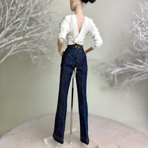 Op de afbeelding: Een pop draagt een witte top met lange mouwen en een lage V-rug en een donkerblauwe spijkerbroek. De broek heeft een hoge taille en een goudkleurige gesp. De pop heeft zwart haar dat in een updo is gestyled.