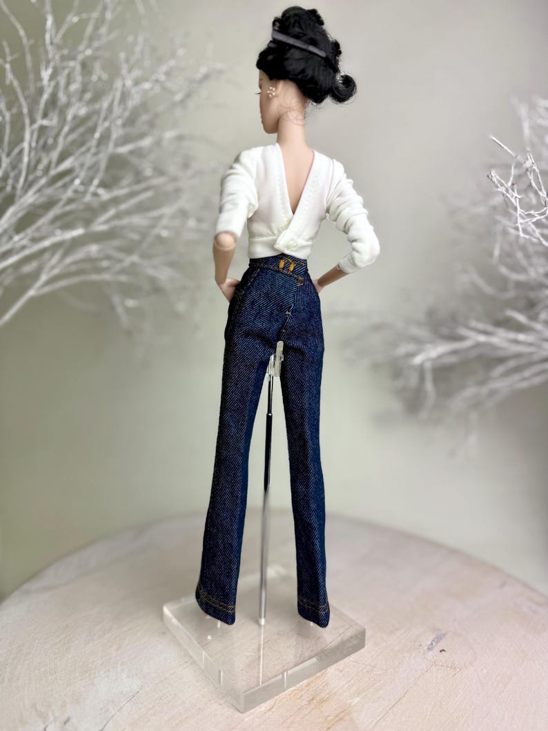 Op de afbeelding: Een pop draagt een witte top met lange mouwen en een lage V-rug en een donkerblauwe spijkerbroek. De broek heeft een hoge taille en een goudkleurige gesp. De pop heeft zwart haar dat in een updo is gestyled.