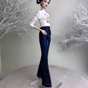 Op de afbeelding: Een modepop met een witte top met bloemenprint en een donkerblauwe flared jeans. De pop heeft donker haar in een knot en roze schoenen. De pop staat op een transparante standaard. De jeans is van donkere denim.