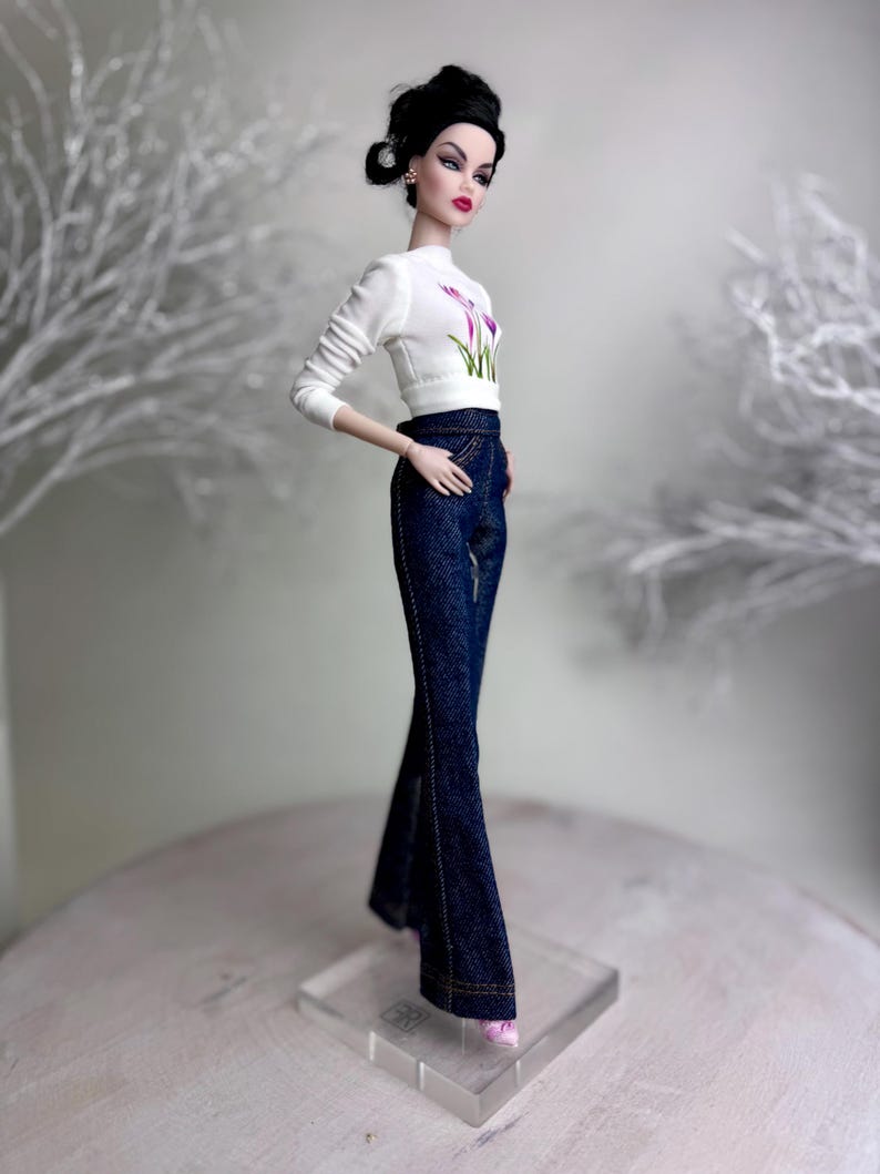 Op de afbeelding: Een modepop met een witte top met bloemenprint en een donkerblauwe flared jeans. De pop heeft donker haar in een knot en roze schoenen. De pop staat op een transparante standaard. De jeans is van donkere denim.