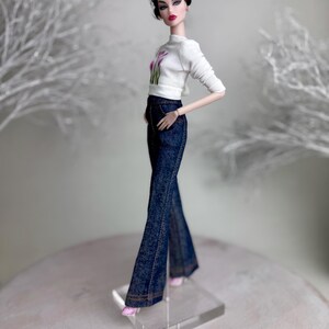 Op de afbeelding: Een modepop draagt een witte top met lange mouwen met een bloemenmotief en een donkerblauwe flared jeans. De pop heeft donker haar en draagt roze schoenen. De pop staat op een transparante standaard.