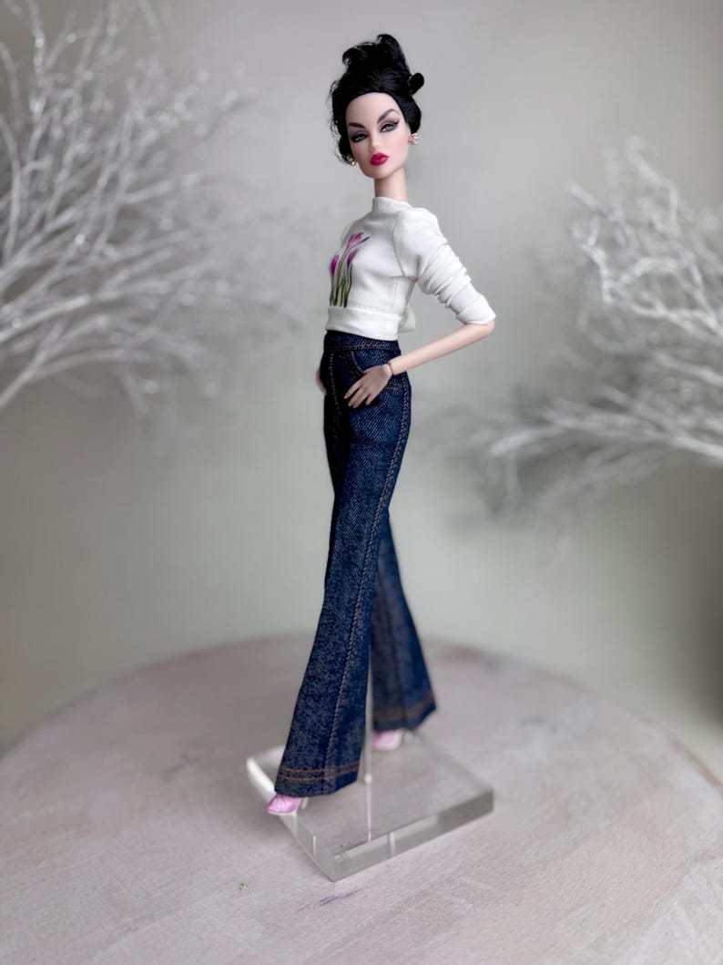 Op de afbeelding: Een modepop draagt een witte top met lange mouwen met een bloemenmotief en een donkerblauwe flared jeans. De pop heeft donker haar en draagt roze schoenen. De pop staat op een transparante standaard.