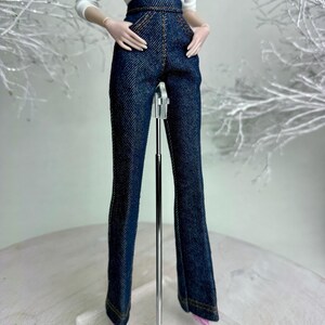 Op de afbeelding: Een pop draagt een witte top met lange mouwen met een bloemenontwerp en een donkerblauwe jeansbroek met hoge taille. De broek heeft een uitlopende pijp en wordt gecombineerd met roze schoenen. De pop staat op een transparante standaard.
