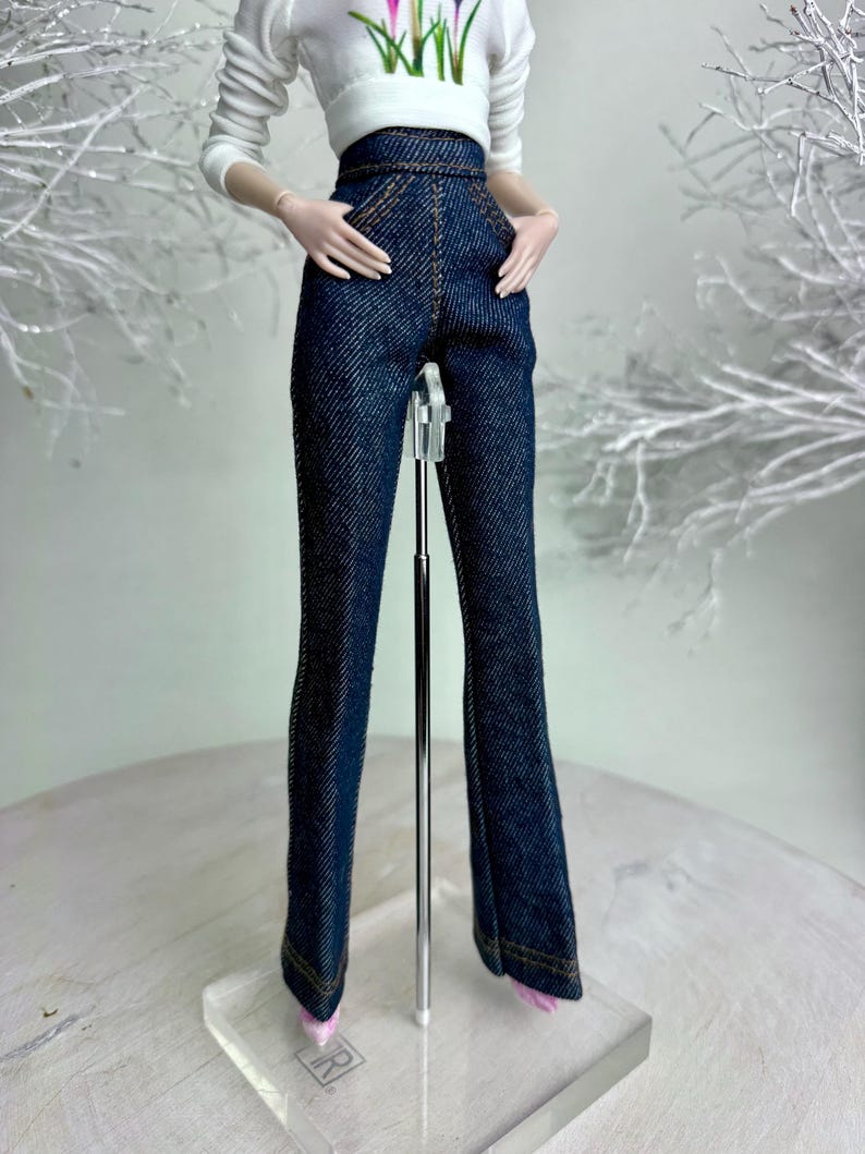 Op de afbeelding: Een pop draagt een witte top met lange mouwen met een bloemenontwerp en een donkerblauwe jeansbroek met hoge taille. De broek heeft een uitlopende pijp en wordt gecombineerd met roze schoenen. De pop staat op een transparante standaard.