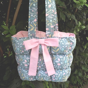 May include: A quilted tote bag featuring a sage green fabric with a multi-colored floral and botanical print. The bag has two matching fabric handles and a large, solid light pink fabric bow attached to the front center. The interior is lined in pink.