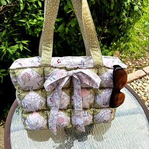 May include: A handmade quilted tote bag with a floral pattern in shades of gray, pink, and green. The bag features a bow and polka dot handles. Sunglasses are tucked into the side. The bag is sitting on a glass table.