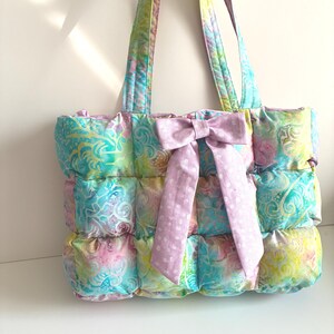May include: A quilted tote bag featuring a multicolored pastel batik pattern in shades of pink, yellow, and blue. The bag has a rectangular, puffed construction with two matching shoulder straps and a large, solid light purple fabric bow.