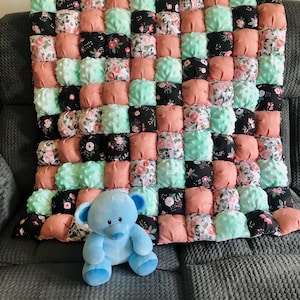 May include: A handmade puff quilt in a grid pattern, featuring squares of coral, mint green, and floral fabrics. A light blue teddy bear sits in front of the quilt on a gray surface.