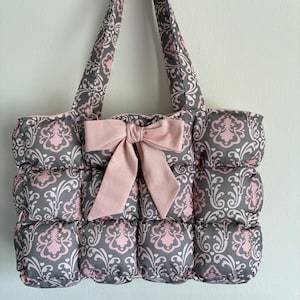 May include: A quilted tote bag featuring a grey and light pink damask pattern. The bag has a puffy, grid-like texture and includes two matching shoulder straps. A large, solid light pink fabric bow is attached to the center front of the bag.
