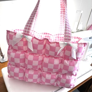 May include: A puffy tote bag featuring a pink and white checkered pattern. The bag has two matching gingham handles and is decorated with two white ribbon bows, each centered with a small pink rose. A ruler is visible at the base of the bag.