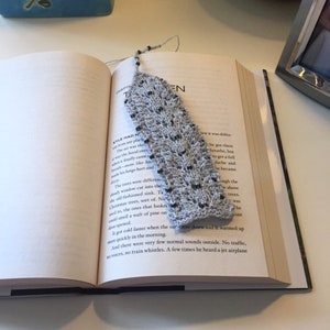 May include: A close-up of a gray and white knitted bookmark with a leaf pattern. The bookmark is laying on top of an open book.