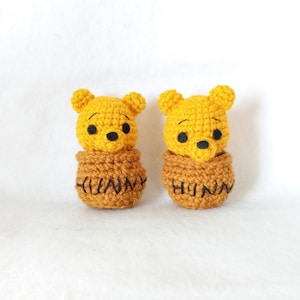 May include: Two handmade crochet figures of Winnie the Pooh. The yellow bear heads sit atop brown honey pots with the word "HUNNY" stitched on the front. The bears have black eyes and noses. The background is white.