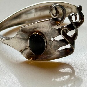May include: A silver bracelet crafted from repurposed silverware. The bracelet features a black oval stone and decorative swirls. The design incorporates the fork tines and handle, creating a unique, handcrafted accessory.