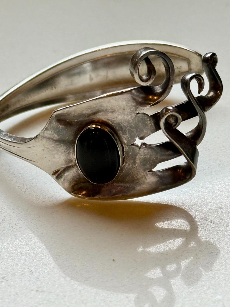 May include: A silver bracelet crafted from repurposed silverware. The bracelet features a black oval stone and decorative swirls. The design incorporates the fork tines and handle, creating a unique, handcrafted accessory.