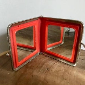 May include: A silver compact mirror with two hinged mirrors. The mirrors have red plastic frames.