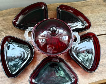 Puede incluir: Un juego de servir de cristal rojo rubí vintage. El juego incluye una fuente para servir cubierta con asas ornamentadas y un pomo decorativo, rodeada de cinco fuentes triangulares. El cristal tiene un acabado brillante.