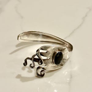May include: A silver-toned ring crafted from repurposed silverware. The ring features a dark oval stone and decorative swirls. The design incorporates the fork's tines and spoon bowl, creating a unique, artistic piece.