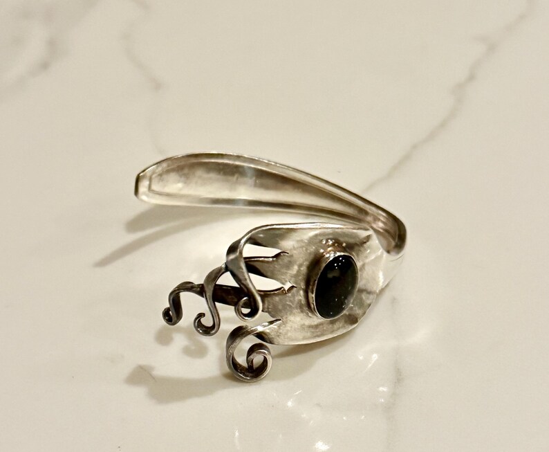 May include: A silver-toned ring crafted from repurposed silverware. The ring features a dark oval stone and decorative swirls. The design incorporates the fork's tines and spoon bowl, creating a unique, artistic piece.