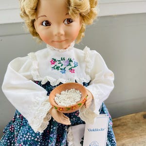 Puede incluir: Muñeca de porcelana con cabello rubio rizado que lleva una blusa blanca con bordados florales y un vestido floral azul y blanco. La muñeca sostiene un pequeño tazón marrón con arroz blanco. La muñeca tiene una etiqueta que dice "Goldilocks" y "Knowles".