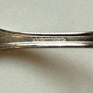 May include: Close-up of a silver utensil with the text "PAT. ARC. 17-17" stamped on the handle. The utensil has a curved design and appears to be made of metal. The background is a light, neutral color.