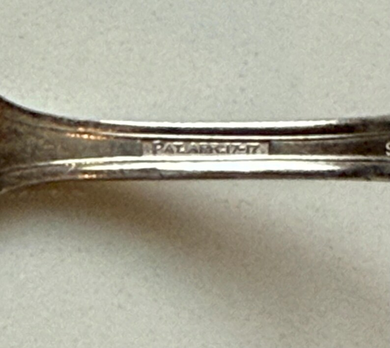May include: Close-up of a silver utensil with the text "PAT. ARC. 17-17" stamped on the handle. The utensil has a curved design and appears to be made of metal. The background is a light, neutral color.