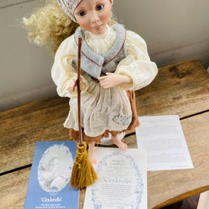 Puede incluir: Una muñeca de porcelana vestida como Cenicienta, sosteniendo una escoba. La muñeca lleva un vestido blanco, un chaleco azul y un pañuelo de flores. La muñeca viene con un certificado de autenticidad.