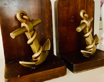 May include: A pair of wooden bookends with gold-colored anchor bookends. The anchors are detailed with rope and have a worn, vintage look.