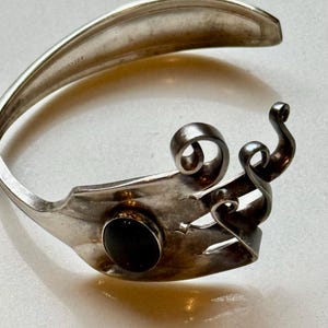 May include: A silver bracelet crafted from a fork, featuring a black stone and decorative swirls. The bracelet has a unique, handcrafted design, showcasing the repurposed silverware. The fork's tines are visible, adding to its distinctive aesthetic.