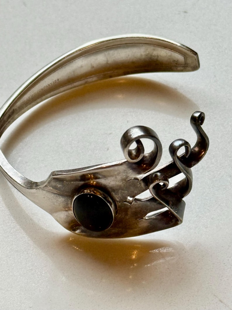 May include: A silver bracelet crafted from a fork, featuring a black stone and decorative swirls. The bracelet has a unique, handcrafted design, showcasing the repurposed silverware. The fork's tines are visible, adding to its distinctive aesthetic.
