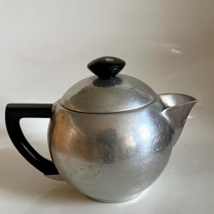 May include: A vintage silver-toned teapot with a rounded body and a black handle and lid knob. The teapot has a spout for pouring and is likely made of metal. The design is simple and classic.