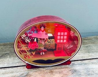 May include: A vintage, oval-shaped, red decorative box with a Japanese theme. Inside, a miniature scene features a geisha, a red lantern, and other traditional elements. The box has a gold-colored trim and a red base.