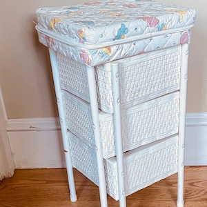 May include: White wicker changing table with three drawers and a padded top. The top is covered in a light blue fabric with a pattern of cartoon characters. The table has a white frame and is set on a wooden floor.