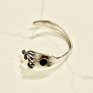 May include: A silver-toned cuff bracelet with a decorative floral design and a dark, round stone. The bracelet is open-ended and rests on a white marble surface. The design incorporates a fork-like shape.