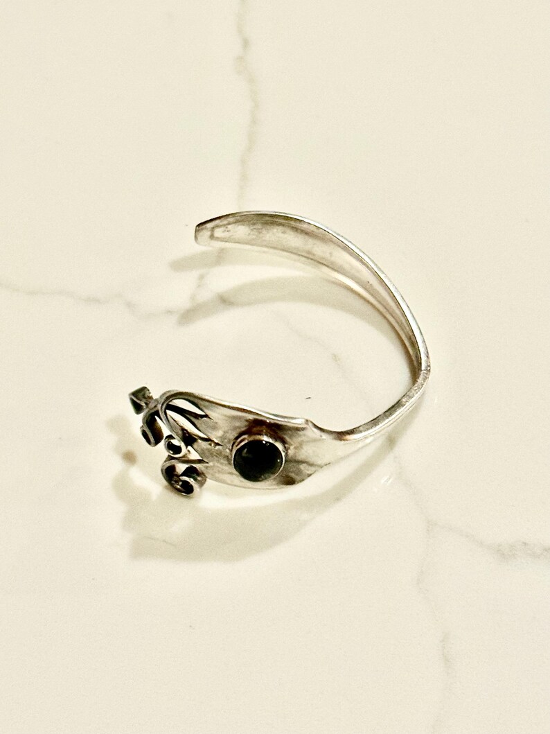 May include: A silver-toned cuff bracelet with a decorative floral design and a dark, round stone. The bracelet is open-ended and rests on a white marble surface. The design incorporates a fork-like shape.