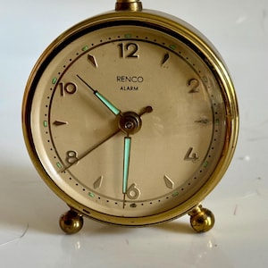 May include: A round gold-tone metal alarm clock with a cream-colored face. The dial features the text RENCO ALARM, gold numerals at 2, 4, 6, 8, 10, and 12, and luminous green-tipped hands. It stands on two small spherical gold-tone metal feet.