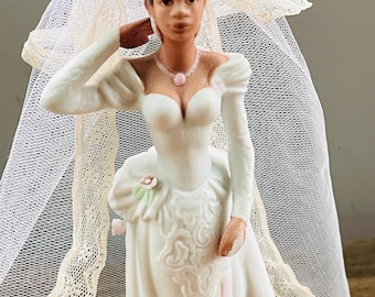 Pode incluir: Estatueta de noiva em porcelana com um vestido de noiva branco com decote em coração e mangas compridas. A noiva usa um véu com acabamento em renda e um colar rosa. A estatueta tem cabelo curto e escuro.