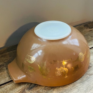 May include: A brown vintage Pyrex mixing bowl with a gold design of corn, chickens, and other farm animals. The bowl has a pouring spout on the side.