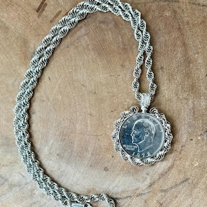 May include: Silver-toned necklace featuring a coin pendant. The pendant has a detailed portrait and the year 1972. The chain is a twisted rope design, and the necklace is displayed on a wooden surface.