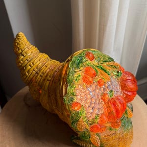 May include: A yellow cornucopia sculpture featuring a woven texture and a collection of molded fruits and vegetables including pumpkins, corn, and grapes. The decorative piece is finished with a glossy sheen and vibrant orange, green, and yellow tones.