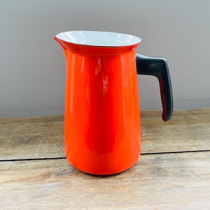 May include: A vibrant orange enamel pitcher with a black handle and a white rim. The pitcher is a classic design, perfect for serving beverages. The pitcher is sitting on a wooden surface.
