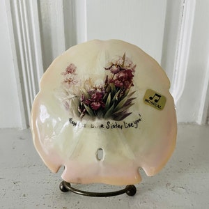 May include: A white seashell with a pink and purple floral design and the text "Happiness is...a Sister Like You!"  The seashell is on a metal stand and has a small gold sticker that says "Musical".