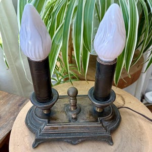 May include: An antique bronze-toned electric candle lamp with two light bulbs. The bulbs are white with a flame-like shape. The lamp has a decorative base and a small switch. The background includes green and white striped plants.