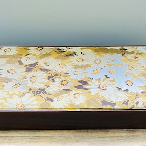 May include: A wooden box with a hinged lid. The lid is decorated with a floral pattern of white daisies on a gold background.