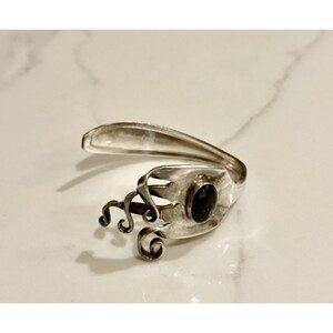 May include: A silver-toned bracelet crafted from a repurposed fork. The design features a dark, oval-shaped stone set within the fork's tines, with decorative swirls. The bracelet is displayed on a white marble surface.