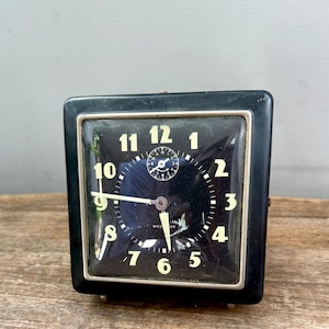 May include: A vintage black square clock with a silver trim. The clock face is black with white numerals and hands. The clock has a small dial at the top center and the word "WESTCLOX" is printed on the face.