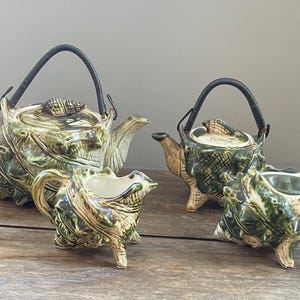 May include: A set of three ceramic teapots shaped like seashells. The teapots are green, brown, and white with a glossy finish. The teapots have black handles and spouts. The teapots are on a wooden surface.
