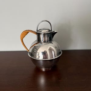 May include: A spherical silver-toned metal pitcher featuring a polished reflective surface, a small spout, a domed lid with a thin metal handle, and a curved side handle wrapped in light brown woven rattan material for a textured grip.